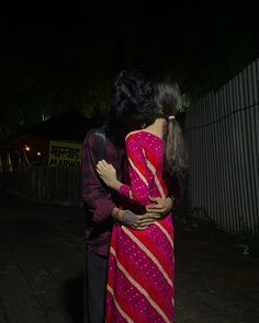 a man and woman standing next to each other on a sidewalk in the dark with their arms around each other