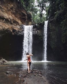 a woman in a bathing suit standing at the base of a waterfall with her arms behind her head