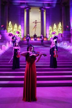 a woman in a red dress playing the violin on stage with other musicians behind her