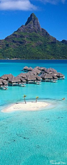 an island in the middle of blue water with huts on it and mountains in the background