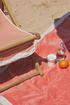 A close-up of a coral beach chair set on the sand, paired with a vibrant beach towel. A water bottle, orange, and enamel cup sit on the towel, capturing a laid-back beach day vibe. Umbrella Chair, Pillow Stack, Beach Blankets, Outdoor Blankets