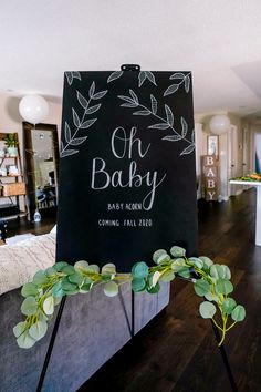 a baby shower sign with greenery on it in the middle of a living room