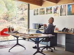 an older man sitting at a desk in front of a large window with pictures on the wall