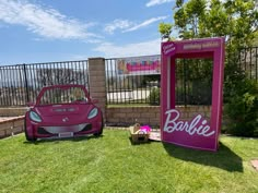 a pink car is parked next to a barbie sign in the grass near a fence