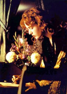 a woman sitting at a table with flowers in front of her and writing on a piece of paper