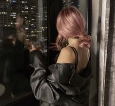 a woman with pink hair standing in front of a window looking out at the city