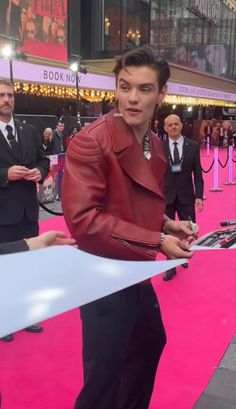 a man in a red leather jacket signing autographs for fans on the pink carpet