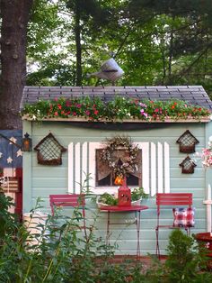 a small blue shed with flowers and potted plants on the windowsill, along with red chairs