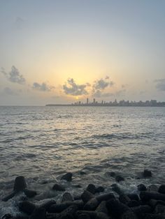 the sun is setting over the ocean with rocks on the shore and buildings in the distance