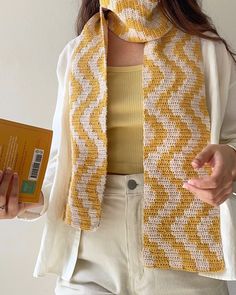 a woman wearing a yellow and white scarf holding an orange book in her hand while standing next to a wall