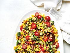 a white plate topped with corn, tomatoes and cucumbers next to a spoon