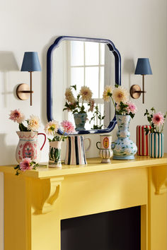 a yellow mantle with vases and flowers on it in front of a large mirror