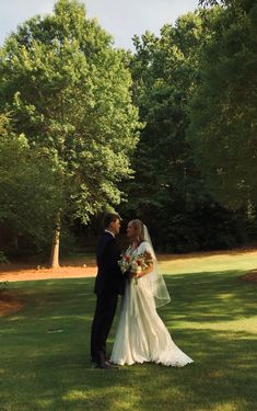 a bride and groom are standing in the grass near some trees on their wedding day