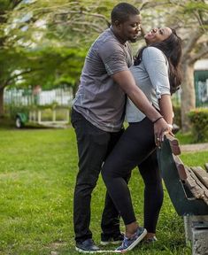 a man and woman standing next to each other on a bench in the grass with trees behind them