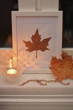 a lit candle sits next to a leaf and bead necklace on a mantle in front of a window