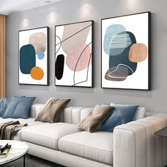 three paintings hang on the wall above a white couch in a living room with two windows