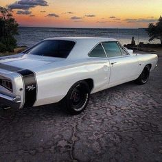 a white muscle car parked in front of the ocean at sunset or sunrise with clouds