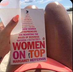 a woman reading a book on the beach with her legs crossed and holding up a drink