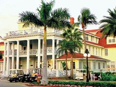 palm trees line the street in front of a large white house with balconies