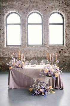 a table with flowers and candles on it in front of three windows, surrounded by chairs