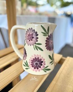 a white coffee mug with purple flowers painted on it sitting on a wooden bench outside