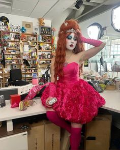 a woman with red hair and makeup is sitting on a desk wearing a pink dress