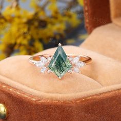 a green and white diamond ring sitting on top of a velvet box with flowers in the background