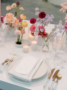 the table is set with white plates, silverware and colorful flowers in vases