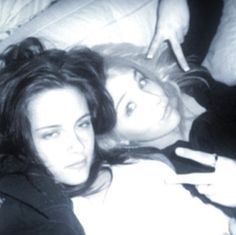 two young women laying in bed with one pointing at the other's ear and holding a cell phone