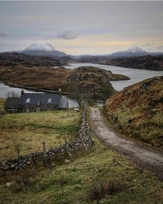 a small house on the side of a dirt road near a body of water with mountains in the background