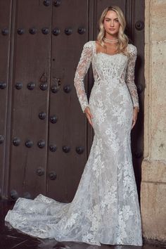 a woman standing in front of a door wearing a white wedding dress with long sleeves