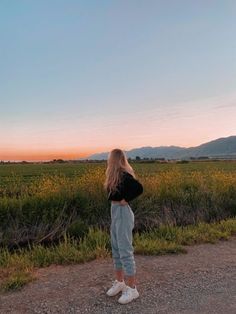 a woman standing in the middle of a field at sunset with her back to the camera