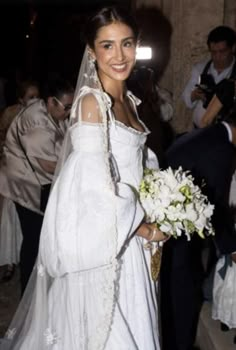 a woman in a white wedding dress holding a bouquet and smiling at the camera while standing next to other people