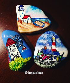 three painted rocks sitting on top of a wooden table next to each other, one has a lighthouse and the other has a bridge