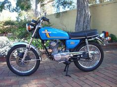 a blue motorcycle parked next to a tree on a brick sidewalk in front of a house