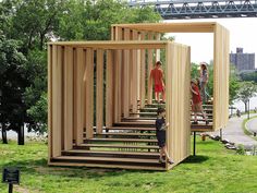 children are standing on wooden structures in the grass near trees and water, while two adults look at them