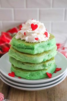 a stack of green pancakes topped with whipped cream and heart shaped candies on a white plate