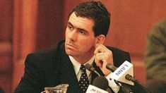 a man in a suit and tie sitting at a table with microphones around him