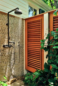 an outdoor shower with wooden shutters next to a window and green plants in the foreground