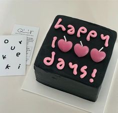 a black cake with pink frosting that says happy birthday on it and three heart shaped candles