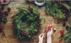 someone decorating christmas wreaths on a wooden table with pine cones and red berries