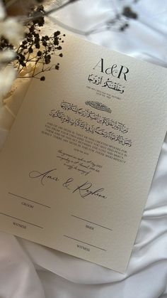 a close up of a wedding card on a bed with white sheets and flowers in the background