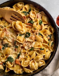 a skillet filled with pasta and spinach on top of a white cloth next to a wooden spoon