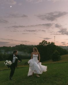 a bride and groom walking in the grass at sunset, with trees in the background