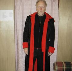 an older man in a red and black robe standing next to a white wall with curtains