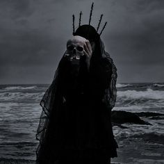 a woman in a black dress holding her face up to the camera while standing by the ocean