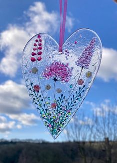 a heart shaped glass ornament hanging from a pink ribbon in front of a blue sky with clouds