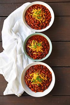 three bowls filled with chili and cheese on top of a white cloth next to a wooden table