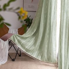 a room with green curtains and a chair in the foreground is a plant that has yellow flowers on it