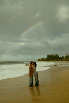 two people standing on the beach with a rainbow in the sky above them and one person holding his arm around the other